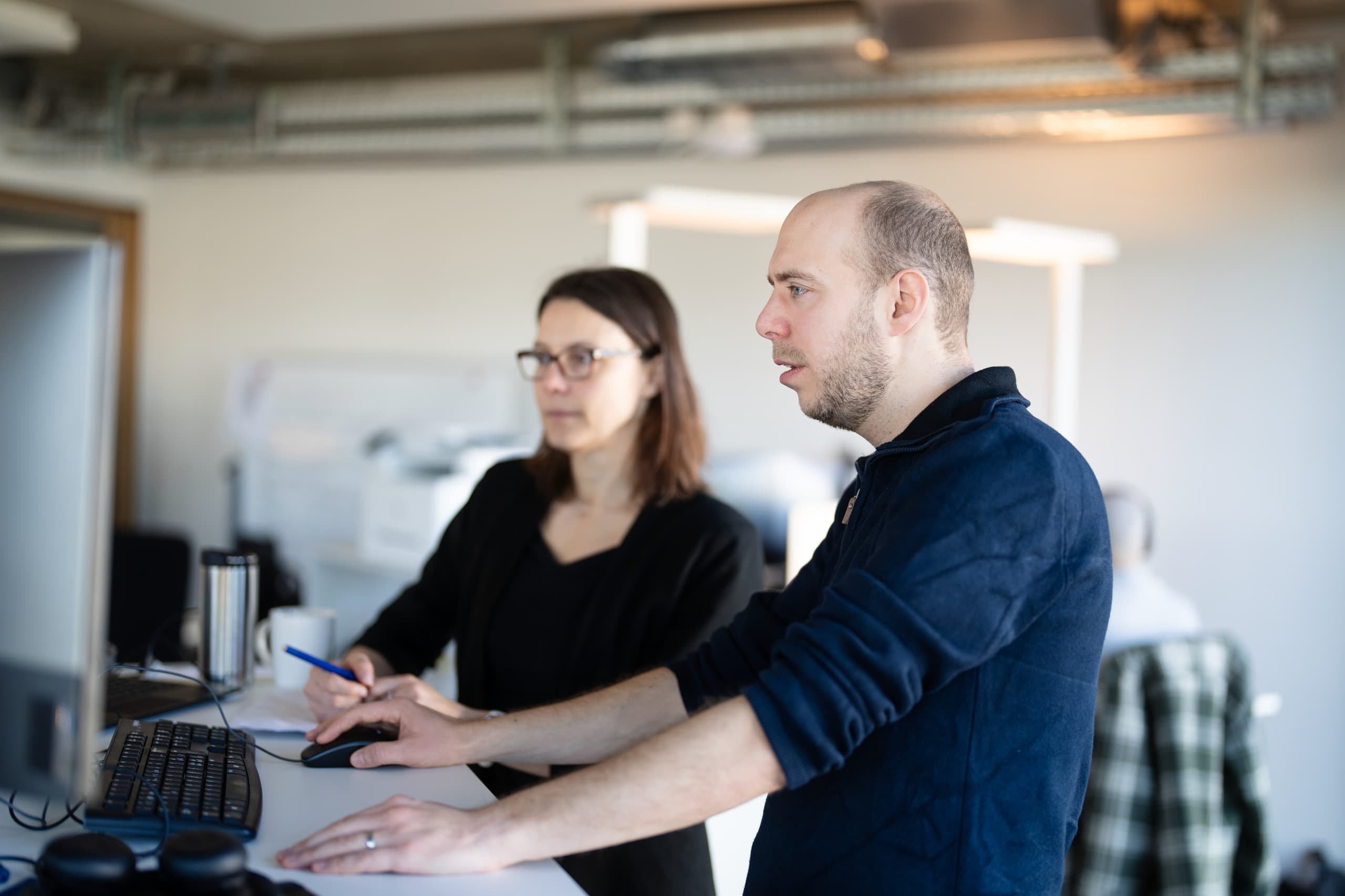 Mitarbeitende arbeiten an Bildschirm-Arbeitsplätzen in einem Büro mit grossen Fenstern und Blick auf die Stadt.